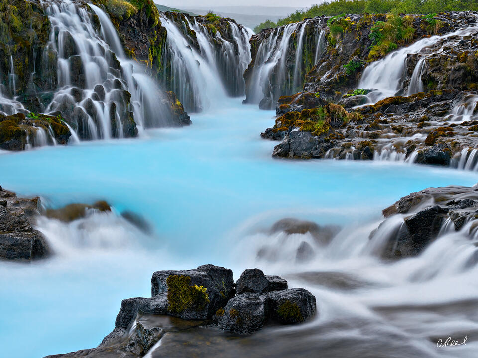 Silky waterfalls cascade over moss-covered volcanic rocks into a vivid turquoise-blue pool in a dramatic Icelandic landscape.