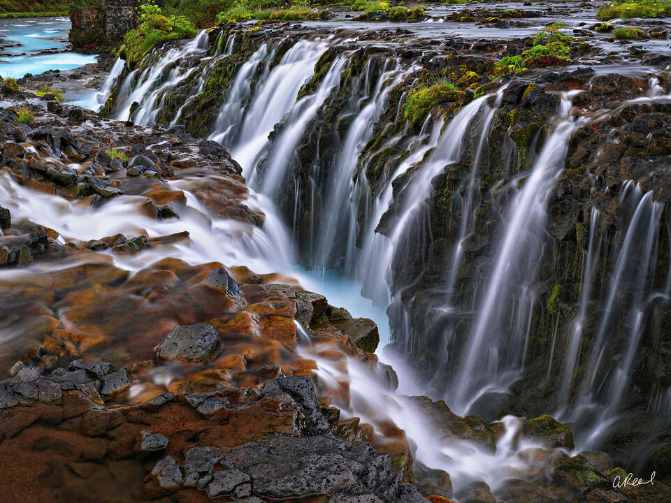 Silky Icelandic waterfalls plunge over dark moss-covered volcanic rock into a deep gorge surrounded by rust-colored stone and green vegetation.