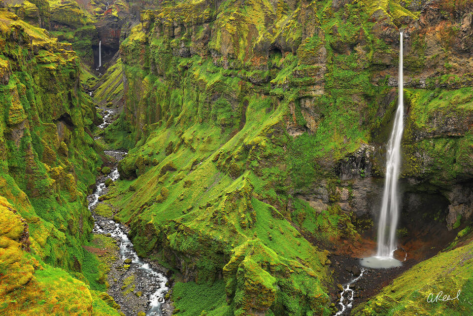 Moss-covered Múlagljúfur Canyon in Iceland with twin waterfalls and a winding stream between steep volcanic cliffs.