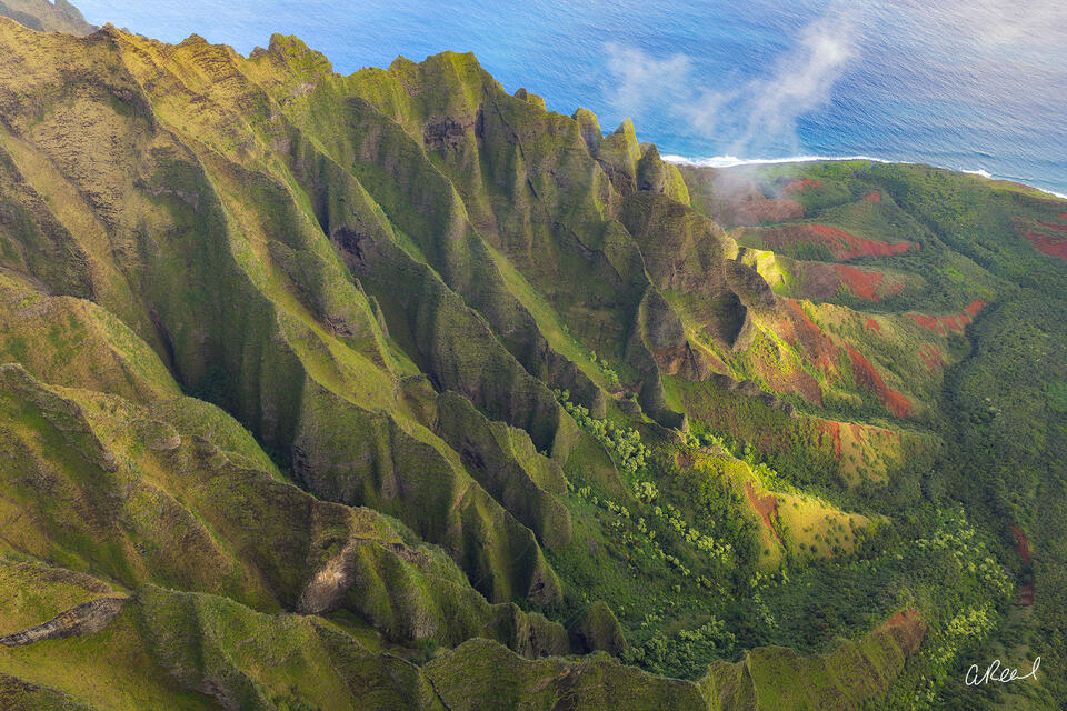 A dramatic aerial photograph of the Nā Pali Coast on Kauai, Hawaii, featuring towering green volcanic cliffs, deep ridges, lush tropical valleys and red earth. 