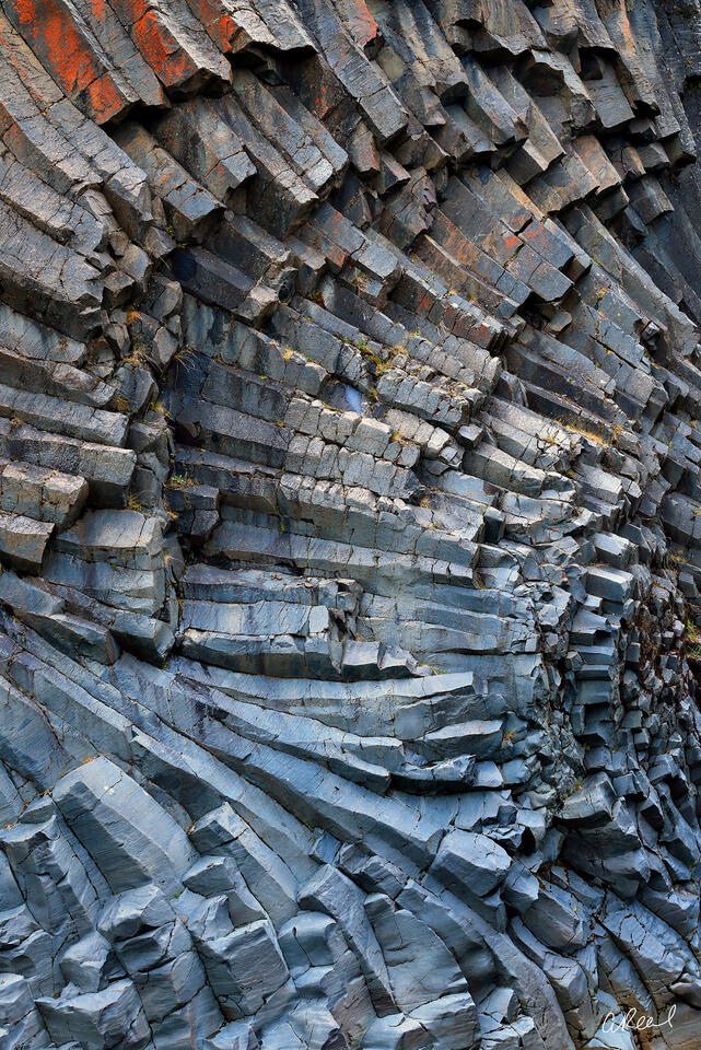 Close-up of a rugged basalt rock wall with fractured, column-like layers in blue-gray tones, streaked with orange lichen and sharp angular textures.