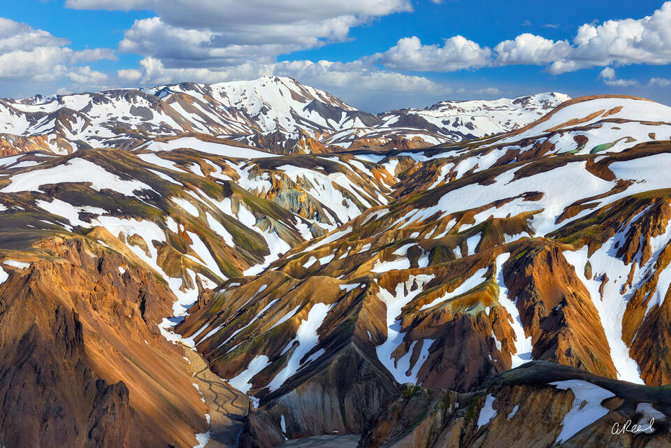 Panoramic landscape photograph of colorful rhyolite mountains and snow-covered valleys in Landmannalaugar, Iceland beneath dramatic summer clouds.