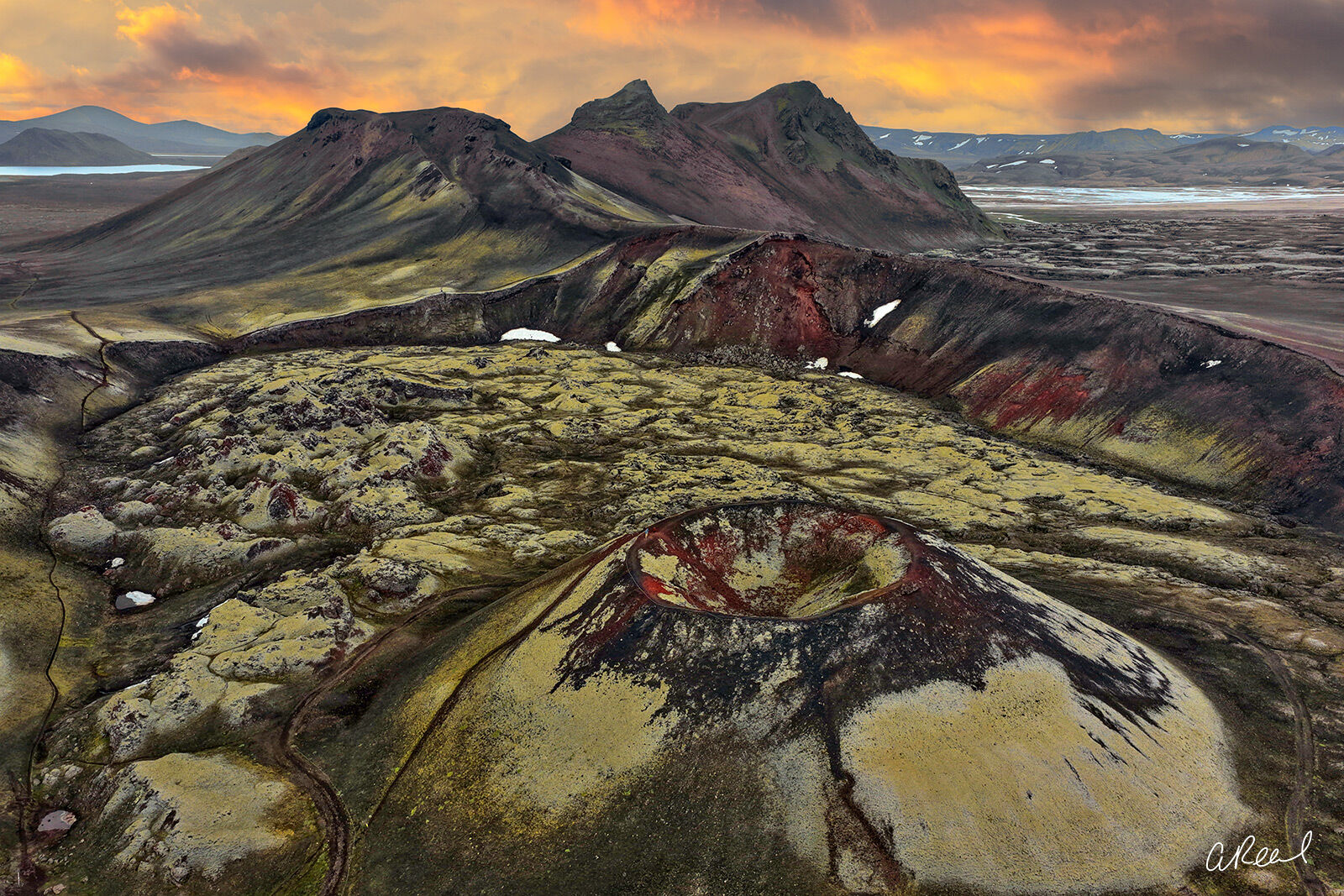 Born From Fire Aerial view of a volcanic crater, moss-covered lava fields and colorful rhyolite mountains beneath a fiery sky in Iceland.